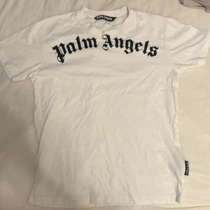 Vit Palm Angels t-shirt med björntryck - Vit t-shirt från Palm Angels med svart logotyp i gotisk stil framtill och ett unikt björntryck på ryggen. Klassisk passform med rund hals och korta ärmar. Tillverkad i mjuk bomull för maximal komfort. Perfekt statement-plagg för dig som gillar streetwear.