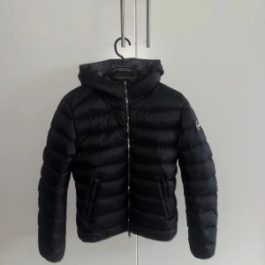 Svart dunjacka från Colmar med huva - NYSKICK! Snygg svart dunjacka från Colmar Originals med huva och quiltad design. Jackan har dragkedja framtill, två fickor med dragkedja och en diskret logga på ärmen. Perfekt för kalla dagar och har en clean, sportig look. 