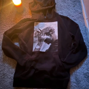 Svart Tupac hoodie Mixtie x Artists storlek S - Svart hoodie med stor Tupac-print på framsidan. Tröjan har huva och en klassisk magficka. Materialet är mjukt och skönt, perfekt för dig som gillar streetwear och vill ha en ikonisk look. Passar dig som vill sticka ut med en cool grafisk design.