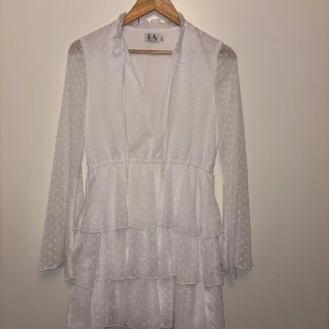 Vit prickig kortklänning med volanger - Superfin vit kortklänning från L.A. med prickigt mönster, v-ringad hals och långa ärmar. Klänningen har flera lager med volanger och tunna snören vid halsen. Perfekt för student eller sommarhäng, riktigt söt och trendig look.