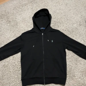 Svart hoodie från Polo Ralph Lauren - Svart hoodie från Polo Ralph Lauren med dragkedja och huva. Tröjan har långa ärmar, snörning vid huvan och klassisk Polo-logga broderad på bröstet. Tillverkad i mjuk bomull och har två fickor framtill. 