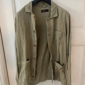 Beige linneskjorta från Morris S - Snygg beige skjorta från Morris i storlek S, tillverkad i linne. Skjortan har klassisk krage, knappar framtill och tre fickor. Perfekt för dig som gillar en chill och stilren look med naturliga material.