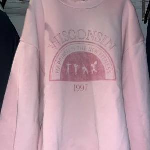 Rosa oversized sweatshirt Wisconsin - Säljer en ljusrosa oversized sweatshirt med tryck 'Wisconsin' och texten 'Happiness is the new fitness 1997' framtill. Tröjan har rund hals och långa ärmar, perfekt för chill dagar. Mjuk och skön känsla, passar dig som gillar streetwear.