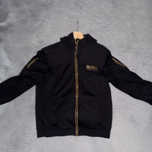  Hugo Boss kofta  - Svart hoodiejacka från Hugo Boss med guldfärgade detaljer och dragkedja. Jackan har huva, långa ärmar och fickor med dragkedja på ärmen. Boss-logga i guld på bröstet och insidan har guldfärgad kant. Perfekt för en clean och stilren look.