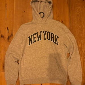 Grå hoodie med New York-tryck - Snygg grå hoodie med stor svartvit New York-text på bröstet. Klassisk modell med huva och känguruficka framtill. Perfekt för dig som gillar streetstyle och vill ha en bekväm och stilren tröja till vardags.
