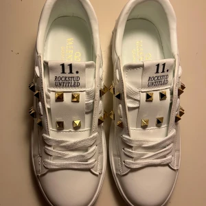 Valentino Rockstud vita sneakers - Snygga vita Valentino Garavani Rockstud Untitled sneakers i skinn med guldiga nitar och coola detaljer. Klassisk rund tå, platt sula och snörning framtill. Perfekta för dig som vill sticka ut med exklusiv streetstyle.