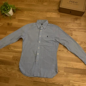 Blå slim fit skjorta från Ralph Lauren - Snygg blå skjorta från Ralph Lauren i slim fit-modell med klassisk krage och knappar framtill. Skjortan har ett subtilt rutigt mönster och den ikoniska broderade Polo-loggan på bröstet. Perfekt för dig som gillar stilrena och tidlösa plagg.