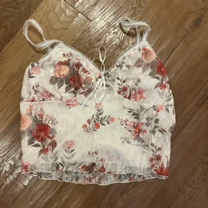 Blommig spets linnetopp med snörning - Säljer en romantisk linnetopp med blommigt mönster i rött och rosa på vit spets. Toppen har smala axelband, söt snörning framtill och genomskinliga spetsdetaljer längs nederkanten. Perfekt för dig som gillar en feminin och trendig stil.