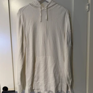 Vit hoodie från C.P. Company - Vit hoodie från C.P. Company med klassisk huva och snörning. Tröjan har en stor ficka framtill och en svart linsdetalj med logga på vänster ärm. Tillverkad i mjuk bomull och har långa ärmar. Perfekt för chill och streetwear.