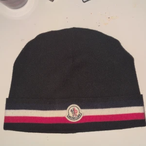 Svart Moncler mössa  - Svart mössa från Moncler med klassiska ränder i marinblått, vitt och rött längs kanten. Broderad Moncler-logga framtill. Tillverkad i en lyxig mix av 70% ull och 30% kaninhår för extra mjuk känsla. Perfekt accessoar för kyliga dagar. Skriv vid frågor, bilder och funderingar (: