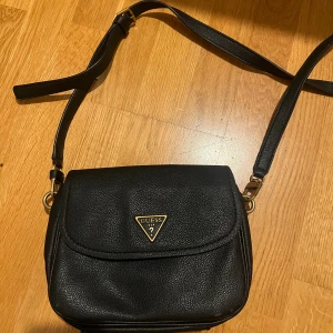 Svart axelväska från Guess - Snygg svart axelväska från Guess med justerbar rem och guldfärgad logga på framsidan. Väskan är tillverkad i strukturerat skinn och har en klassisk, stilren design med lock. Perfekt storlek för det viktigaste och passar till många olika outfits.