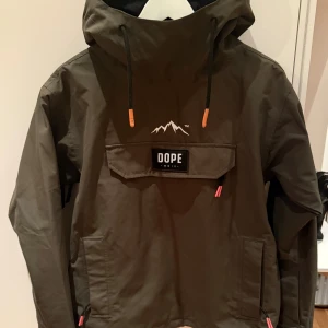 Olivgrön skidjacka från Dope XXS - Säljer en olivgrön jacka (modell Blizzard) från Dope med stor huva och ventilation under ärmarna. Jackan har snörning i huvan och är vatten och vindtålig, stor ficka med Dope-logga och orange detaljer på snören. Materialet är slitstark återvunnen polyester och jackan har långa ärmar med kardborre vid mudden.