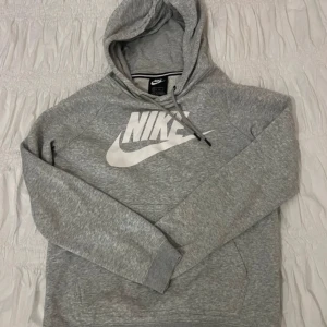 Grå hoodie från Nike med logga - Säljer en grå hoodie från Nike i storlek XS. Tröjan har en stor vit Nike-logga på bröstet, huva med snörning och en klassisk magficka. Perfekt för chill dagar eller när du vill ha en sportig look. Mjuk och skön med lång ärm.