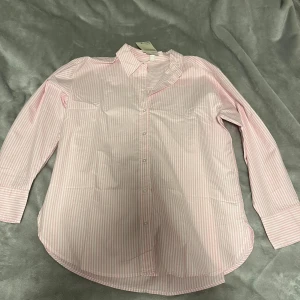 Rosa och vit randig skjorta H&M - Snygg skjorta från H&M i storlek M med rosa och vita smala ränder. Klassisk krage, långa ärmar och knappar hela vägen framtill. Tillverkad av bomull med 20% återvunnen bomull. Perfekt för dig som gillar stilrena och fräscha plagg.