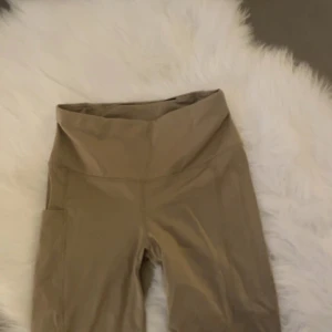 Beige leggings med hög midja XS/S - Snygga beige leggings med hög midja och praktisk ficka på sidan. Perfekta för dig som gillar en clean och enkel look. Stretchigt material som sitter skönt och följer kroppen. Passar till träning eller chill.