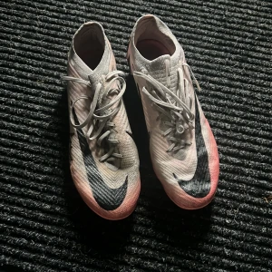 Nike mercurial vapor 15 elite - Säljer ett par snygga Nike fotbollsskor i ljusrosa med svarta detaljer och stor Nike-logga på sidan. Skorna har snörning och är tillverkade i syntetmaterial med en smidig och sportig form. Perfekta för dig som vill ha stil på planen.
