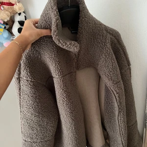 Brun teddyjacka från Gina Tricot - Mysig brun teddyjacka från Gina Tricot i storlek M. Jackan har hög krage, rak passform och dragkedja framtill. Materialet är fluffigt och fleece-liknande, perfekt för kyliga dagar. Enkel design utan mönster eller extra detaljer. Knappt använt, för stor.