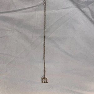 Guldigt halsband med bokstavshänge - Snyggt halsband i guldton med ett hänge i form av bokstaven 'm' dekorerat med glittriga stenar. Perfekt för dig som vill ha ett personligt smycke med lite extra bling. Kedjan är tunn och stilren, passar till många olika looks.