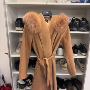 Beige kappa med päls och bälte - Snygg beige kappa från Hollies med stor huva och fluffig fuskpäls runt kragen. Kappan har knytbälte i midjan och långa ärmar, perfekt för kyliga dagar. Stilren och trendig modell som ger en lyxig känsla. Bra skick 