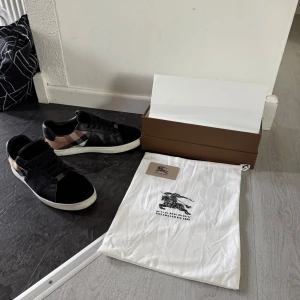 Burberry svarta sneakers med rutig detalj - Snygga svarta sneakers från Burberry med klassisk rutig panel i beige, svart och vit på sidan. Skorna har snörning, rund tå och vit platt sula. Materialmix av skinn och mocka ger en lyxig känsla. Perfekta för dig som gillar designer och vill sticka ut med stil. Använda i några få månader och är i utmärkt skick.