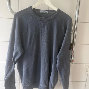 Grå/blå sweatshirt från Stone Island - Grå sweatshirt från Stone Island med klassisk rund hals och ribbade muddar. Tröjan har den ikoniska loggan på vänster ärm och är tillverkad i mjuk bomull. Perfekt för dig som gillar stilrena och avslappnade plagg. Sparsamt använd, mycket gott skick 