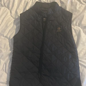 Svart quiltad dunväst från Morris - Quiltad svart dunväst från Morris med guldfärgad logga på bröstet. Västen har ståkrage och dragkedja framtill. Snyggt rutmönster över hela västen och en stilren look som funkar till många outfits. Perfekt för dig som gillar klassisk stil med en modern twist.