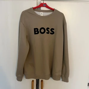 Beige sweatshirt med BOSS-tryck - Snygg beige sweatshirt från BOSS med svart tryck framtill. Klassisk rund halsringning och långa ärmar. Perfekt för dig som vill ha en enkel men stilren tröja med statement. Tillverkad i mjuk bomull för maximal komfort.