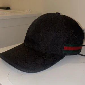 Svart Gucci keps med logomönster - Snygg svart keps från Gucci med diskret GG-logomönster över hela ytan. Klassisk böjd skärm och justerbar rem bak i skinn. Sidan har den ikoniska gröna och röda randen. Materialet är bomull och skinn, perfekt för dig som vill ha en lyxig streetstil.