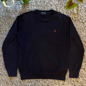 Mörkblå tröja från Polo Ralph Lauren - Klassisk mörkblå tröja från Polo Ralph Lauren med rund halsringning och röd broderad logga på bröstet. Tröjan är långärmad och har ribbade muddar vid ärmslut och nederkant. Perfekt för en stilren och enkel look.
