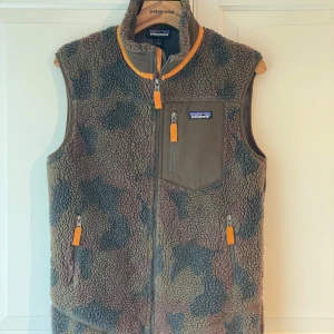Patagonia fleece Retro-X väst - Vindtät fleeceväst som kombinerar snygg  retrostil med teknisk funktionalitet.  Mycket fint skick. Köpt på Patagonia, har mest hängt i garderoben.  Färg: diskret camo Tillverkad i varm ¼"-pile fleece med vindavvisande membran och mjuk meshfodrad insida som transporterar bort fukt. Integrerad barriär som stoppar kalla vindar Tjock fleece som isolerar i ruggigt väder Bröstficka med dragkedja + två fodrade handfickor Passform, sitter bra på mig som är 177cm. 