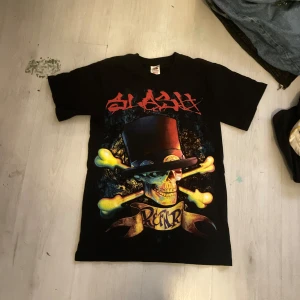 Slash Tour 2010 t-shirt svart S - Grym svart t-shirt från Slash Tour 2010 med fet döskalle och korslagda ben på framsidan, coolt tryck i rött och gult. Baksidan har stor röd text och döskallemotiv. Klassisk rund hals och korta ärmar. Materialet är tung bomull, perfekt för dig som gillar rock och snygga bandtees.