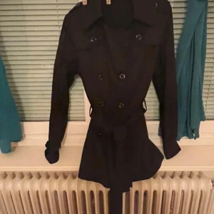 Svart trenchcoat från Vila, storlek L L - Snygg svart trenchcoat från Vila i storlek L. Jackan har dubbelknäppning, klassisk krage och bälte i midjan för en schysst siluett. Långa ärmar och stilrena knappar framtill. Perfekt för dig som vill ha en tidlös och clean look.