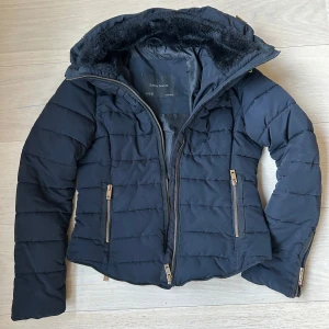Zara jacka - Marinblå pufferjacka från Zara med gömd huva och snygga guldfärgade dragkedjor. Jackan är quiltad och har två fickor framtill samt dragkedjor vid ärmsluten. Perfekt för kyliga dagar och har en stilren, figurnära passform.