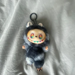 Labubu secret - Säljer en unik och fluffig nyckelringsväska från Pop Mart. Väskan är formad som en söt figur med regnbågsfärgat ansikte och päls i mörkgrå och ljusgrå toner. Perfekt att hänga på väskan eller använda som liten accessoar.