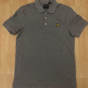 Grå pikétröja från Lyle & Scott - Snygg grå pikétröja från Lyle & Scott med klassisk krage och tre knappar framtill. Tröjan har det ikoniska gula logobroderiet på bröstet och är tillverkad i mjuk bomull. Perfekt för en clean och stilren look.