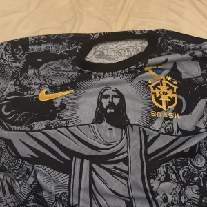 Brasil Nike fotbollströja svart/grå - Unik Brasilien fotbollströja från Nike med stort tryck av Kristusstatyn och detaljerade grafiska mönster. Svart och grå bas med gula detaljer, CBF-emblem och Nike-logga på bröstet. perfekt för match eller streetstyle.