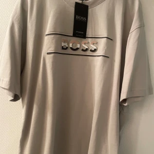Beige t-shirt från BOSS  - Snygg beige t-shirt från BOSS med stor logga framtill i 3D-tryck och raka linjer. Klassisk rund hals och korta ärmar. Tillverkad i mjuk bomull som känns skön mot huden. Perfekt för en clean och stilren look.