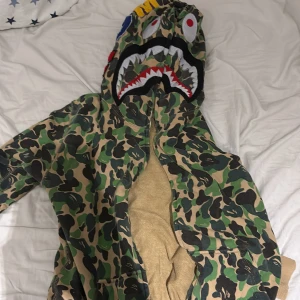 BAPE camo hoodie med shark-tryck - Säljer en ikonisk BAPE hoodie i klassiskt grönt camo-mönster med hel dragkedja och haj-ansikte på huvan. Hoodien har broderade detaljer, ribbade muddar och är tillverkad i mjuk bomull. Perfekt statement-plagg för streetwear-entusiasten.