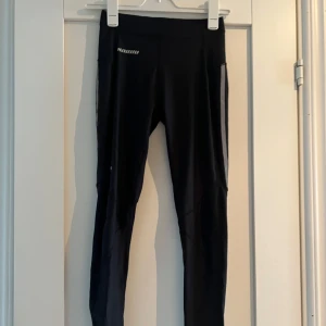 Svarta löpartights  - Svarta löpartights  med diskret logga på låret och liten dragkedjeficka bak. Tightsen är tighta och har hög midja, perfekta för träning. Tillverkade i stretchigt syntetmaterial för maximal rörelsefrihet