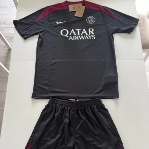 PSG träningsset svart vinröd - Snyggt PSG träningsset från Nike i svart med vinröda axel- och sidopaneler. Tröjan har korta ärmar, Paris Saint-Germain-logga och Qatar Airways-tryck på bröstet. Tillhörande shorts med klubbmärke och Nike-logga. Tillverkat i lätt och ventilerande polyester.