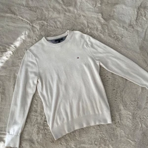 Vit stickad tröja Tommy Hilfiger  - Vit stickad tröja från Tommy Hilfiger i storlek S. Klassisk rund halsringning och diskret broderad logga på bröstet. Tröjan har långa ärmar och ribbade muddar för en clean och stilren look. Perfekt för lager-på-lager under hösten och vintern.