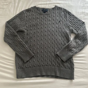 Grå kabelstickad tröja Polo Ralph Lauren - Kabelstickad grå tröja från Polo Ralph Lauren i klassisk modell med rund halsringning och ribbade muddar.  Storlek XL från 18 - 20 år så den sitter som M/L. Skriv vid intresse!