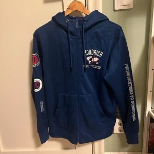 Blå hoodie från Hoodrich med tryck - Blå hoodie från Hoodrich med vita tryckdetaljer på bröstet, ärmarna och huvan. Tröjan har dragkedja framtill, snörning i huvan och känguruficka. Snygga patches på ena ärmen och texten 'From Nothing To Something' på andra ärmen. Perfekt streetwear-vibe.