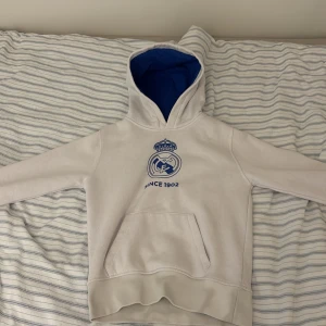 Vit Real Madrid hoodie med blått foder - Säljer en vit hoodie från Real Madrid med klubbens logga och texten 'Since 1902' i blått framtill. Tröjan har blått foder i huvan, känguruficka och långa ärmar. Perfekt för dig som älskar fotboll och vill visa ditt stöd för laget. Kvalité 9,6/10. Används 3-5 gånger 