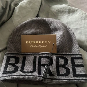 Grå stickad mössa från Burberry - Snygg grå stickad mössa från Burberry med bred uppvikt kant och stor svart logotyp runtom. Klassisk och stilren design som passar perfekt till kalla dagar. Tillverkad i mjukt material för extra komfort och värme. (Aldrig använd)