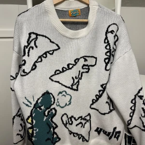 Vit stickad tröja med dinosaurier  - Super gullig och mysig stickad tröja med dinosaurier! Köpte på nätet och osäkert på märket men den är knappt använd. Sänkt pris vid snabb affär!