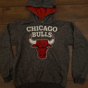 Chicago Bulls grå hoodie med tryck - Grå hoodie med Chicago Bulls-logga och stor röd tjur på bröstet. Mjuk insida, röd huva på insidan och klassisk känguruficka framtill. Perfekt för dig som gillar basket och streetstyle.