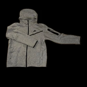 Grå Nike hoodie med dragkedja L - Snygg grå hoodie från Nike i storlek L. Tröjan har hel dragkedja, huva och svarta detaljer vid fickor och ärmar. Perfekt för chill eller träning, med lång ärm och sportig look. Materialet är mjukt och skönt, bomull och polyester.