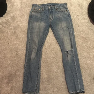 Levi's 501 ljusblå jeans W30 L32 - Klassiska Levi's 501 jeans i ljusblå tvätt med raka ben och knappgylf. Jeansen har en slitning på ena knät och tydliga kontrastsömmar. Tillverkade i bomull och har den ikoniska läderpatchen bak i midjan.