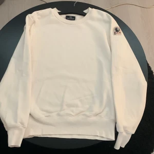 Vit fleece-tröja från Parajumpers L - Vit fleece-tröja från Parajumpers i storlek L. Klassisk rund halsringning, ribbade muddar och en snygg broderad patch på ärmen. Perfekt för dig som gillar stilren och clean look med en sportig touch. Nästan helt ny bara använt den 1 gång och den är tvättad 1 gång. Köpt på season i Jönköping 